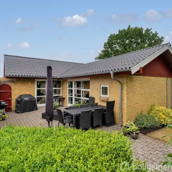 Gulstenshus med gråt tag ligger i en have med flisebelagt terrasse. Der er sorte havemøbler og en grill, omgivet af grønne buske og træer under en skyet himmel.