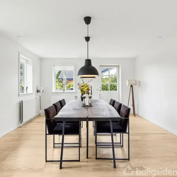 Spisebord med otte stole står i en lys spisestue. En stor lampe hænger over bordet, og der er vinduer mod en have. Gulvet er træ, og væggene er hvide.