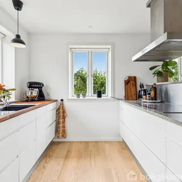 Et lyst, moderne køkken med hvide skabe og trægulve. Køkkenmaskiner og krydderurter er placeret på bordplader af træ og granit. Store vinduer giver udsigt til grønne omgivelser.