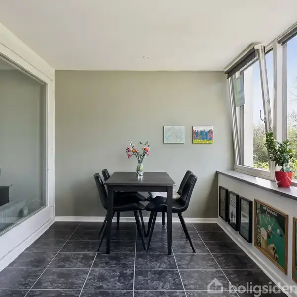 Et bord med fire stole står i et lyst rum. Bordet har en vase med blomster. Til venstre ses en åben skydedør til et kontor, og vinduer til højre giver udsigt til natur.