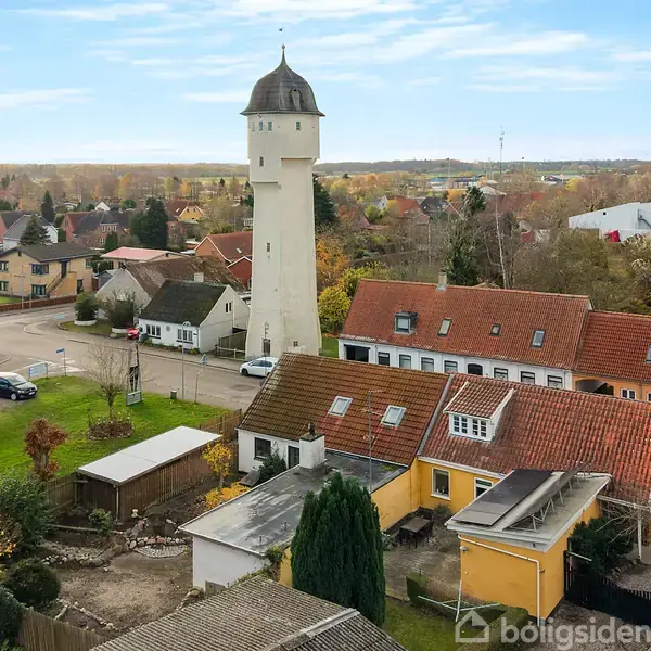 Vandtårn står højt blandt huse med røde tage, placeret i en villaby med træer i efterårsfarver i baggrunden under en blå himmel.
