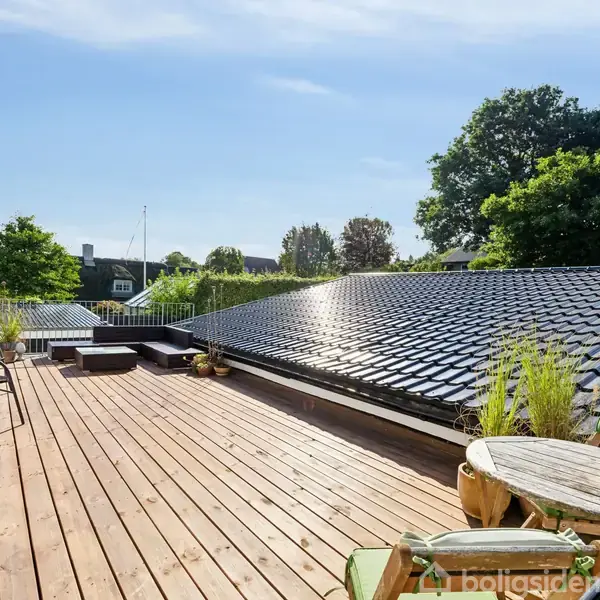 En træterrasse med stole og planter ligger ved siden af et sort tegltag, omgivet af grønne træer og himmel med lette skyer, i et boligområde.