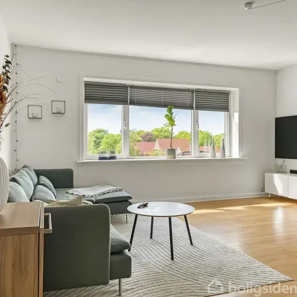 Stue med grøn sofa og runde borde står på et tæppe. En stor vase med tørrede blomster pynter. Store vinduer slipper lys ind, og en trappe ses i højre hjørne.