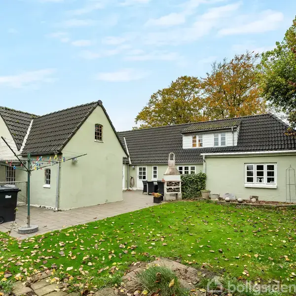 Et grønt hus med mørkt tag står i en have med græsplæne og flisebelagt gårdhave. Der er skraldespande og et udendørs pejs i baghaven, omgivet af træer.