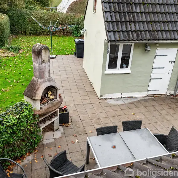 Terrassebord med stole står på flisebelagt terrasse. Grillovn ved siden af grøn græsplæne. Lille, hvid bygning i baggrunden med sort tag. Ingen tekst på billedet.