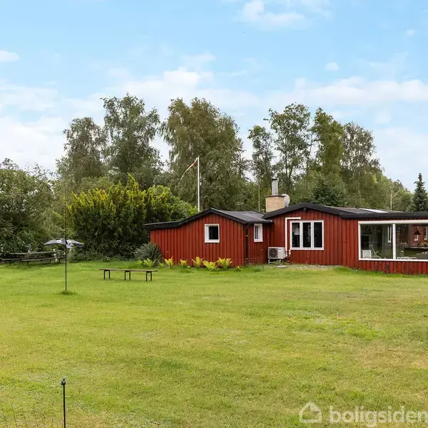 Et rødt sommerhus står stille på en stor græsplæne, omgivet af træer under en blå himmel. Et bord og parasol ses til venstre for huset.