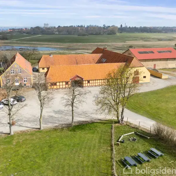 En gård med orange tage ligger blandt grønne marker. Biler holder parkeret foran bygningerne. I baggrunden ses en flod og en by i horisonten.