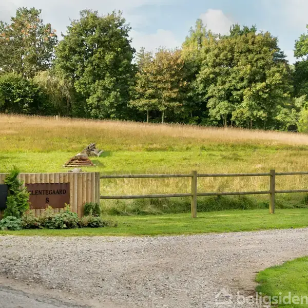 Skilt angiver "VENTEGÅRD" ved hegn. Grusvej løber forbi, grøn mark med træer i baggrunden skaber en landlig atmosfære.