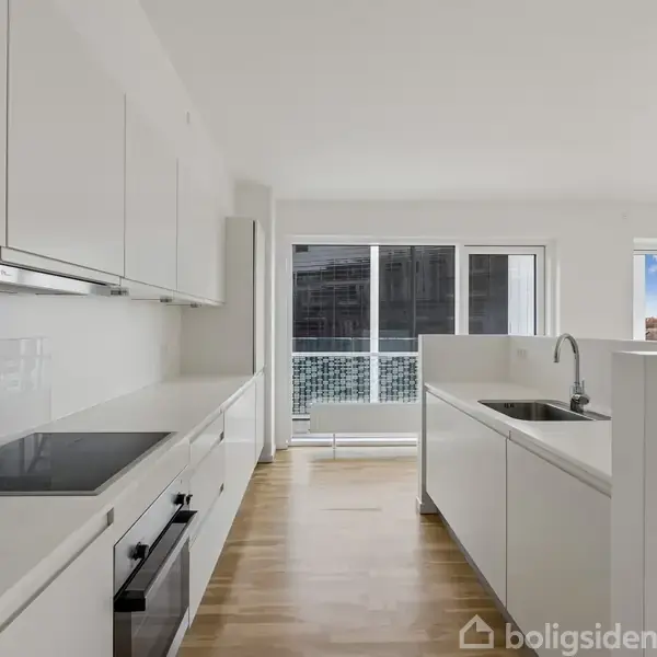 Et moderne køkken med hvide skabe og indbygget kogeplade står i en lys lejlighed med trægulve. I baggrunden ses store vinduer, der leder til en balkon.