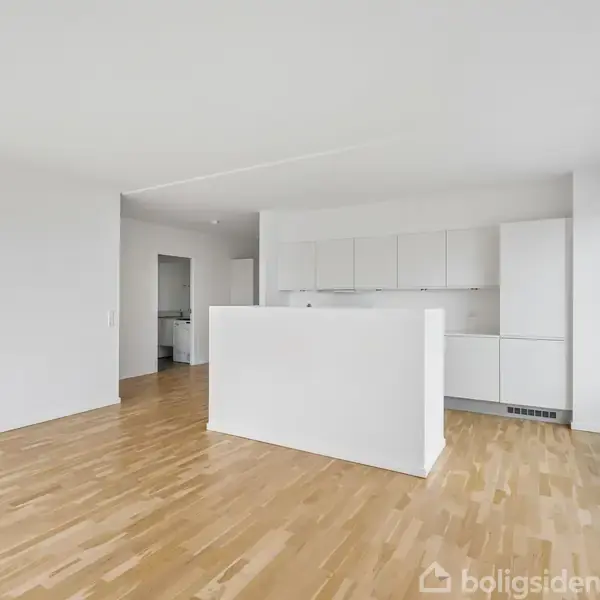 Et moderne køkken med hvide skabe og en ø, uden apparater. Trægulve strækker sig gennem rummet, som har store vinduer til højre, der giver naturligt lys.