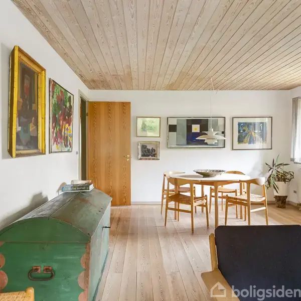 Et spisebord med fire stole står i et lyst rum med trægulv. Væggen er dekoreret med flere farverige malerier. En stor grøn kiste og en plante er også til stede.