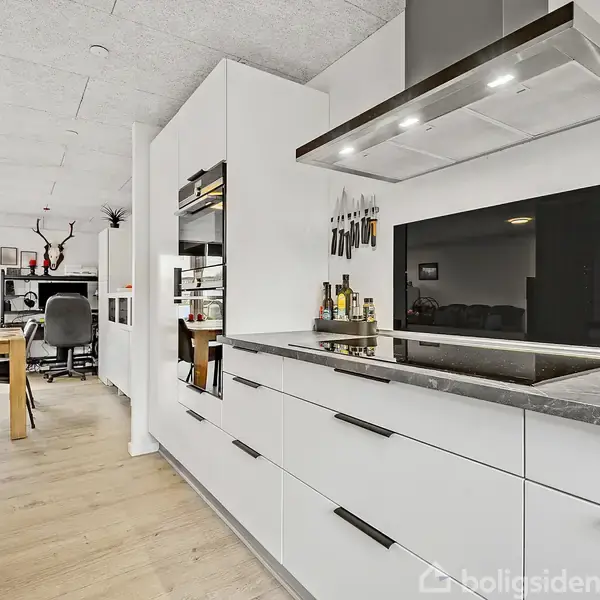 Et moderne køkken med hvide skabe og en sort komfurplade. Knive hænger på væggen. I baggrunden er et spisebord med stole og en stuesektion med en kontorstol.