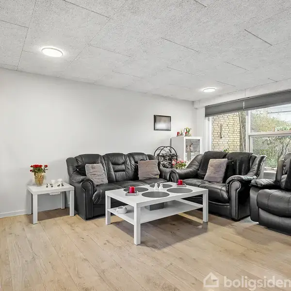 Sort lædersofa står i stuen med et sofabord foran; rummet har lyse trægulve, hvide vægge og store vinduer, der åbner op til en have. Røde lysestager pynter.