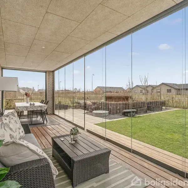 Overdækket terrasse med sofa og bord, set ud mod haven gennem glaspartier. Udenfor ses et grønt græsareal og nabohuse i baggrunden. Vandret træbeklædning pryder gulvet.