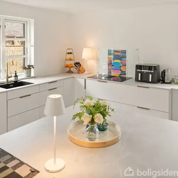 Et moderne køkken med hvide skabe. Bordet har en lampe og en vase med blomster. Vinduer viser en have udenfor. Apparater som brødrister og kaffemaskine står på bordpladen.