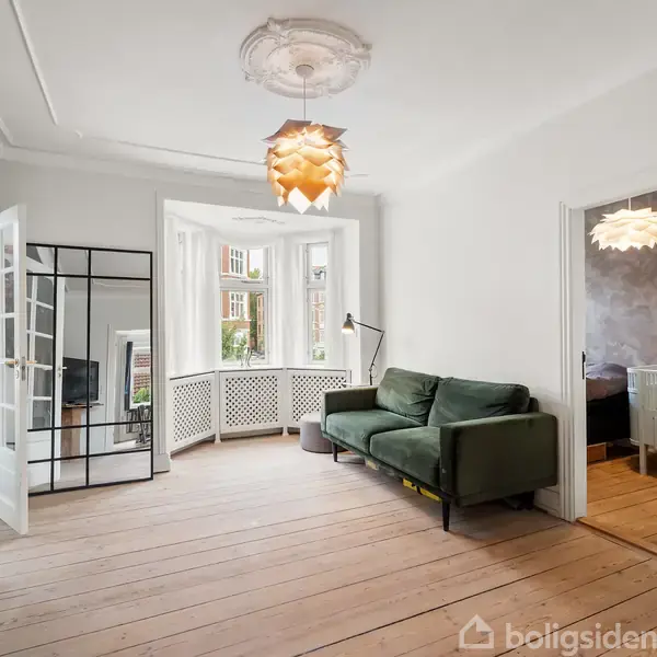 Grøn sofa står i en lys stue ved åbent vindue. En unik loftslampe hænger over trægulvet. Til højre ses en dør til et børneværelse.