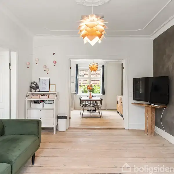 Grøn sofa står i en stue med lyse trægulve og moderne loftslampe. En fladskærms-TV hænger på en væg ved siden af et åbent rum med spisebord og stole.
