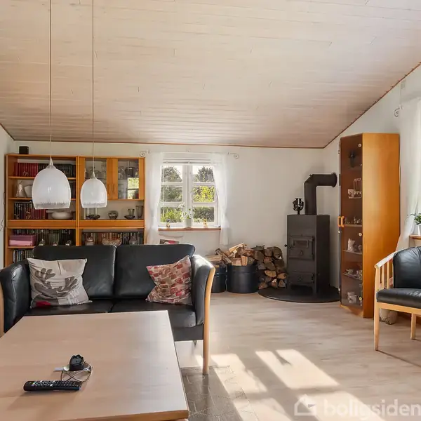 Sort lædersofa og lænestole står i en stue, omgivet af en brændeovn og reol. Rummet har lyse vægge og et træbord med fjernbetjening. Store lysindfald gennem vinduerne.