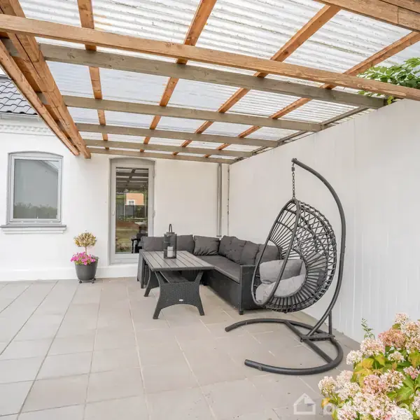 Terrasse med hængestol og sofagruppe under trætagskonstruktion. Grå fliser dækker gulvet, mens planter i potter og blomsterdekorationer pynter omgivelserne op mod hvid husvæg.
