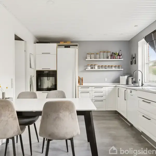 Køkkenbordet med fire beige stole står i en moderne, hvidt køkken med komfur, opvaskemaskine og åbne hylder fyldt med krydderier. Væggene har indrammet kunst.