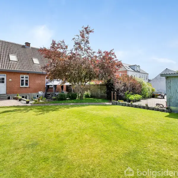 Rødt murstenshus med sort tag står stille i en solrig have. Omkranset af velholdt græsplæne og frodig hæk, ved siden af et træ i blomst. I baggrunden ses en garage.