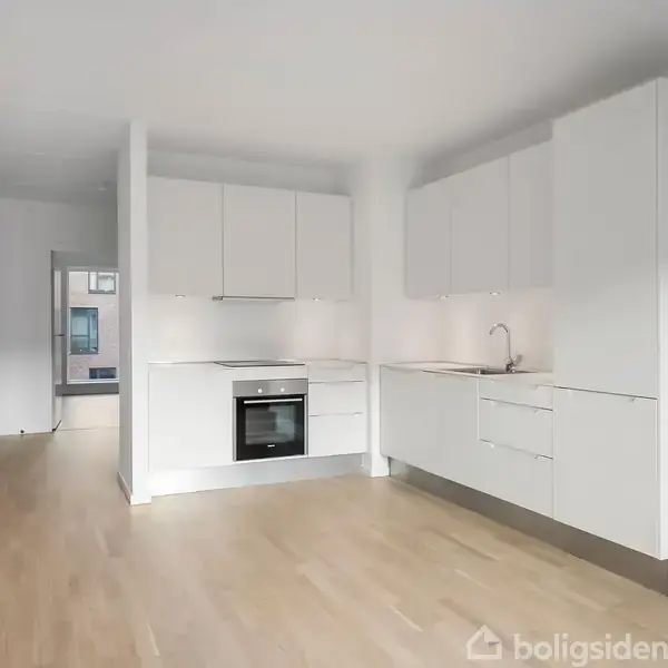 Et lyst, åbent rum med træparketgulv indeholder et moderne, hvidt køkken i venstre side. I baggrunden fører en glasdør ud til en balkon med udsigt til en bygning.