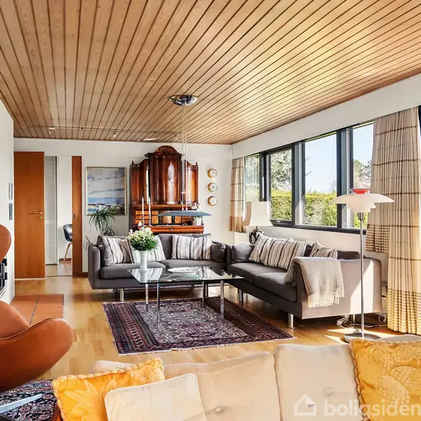 Brunt lænestol står i en stue med et moderne design. Sort sofa og sofabord på tæppe. Store vinduer giver udsigt til en have. Hylder med dekorationer. Ingen tekst synlig.
