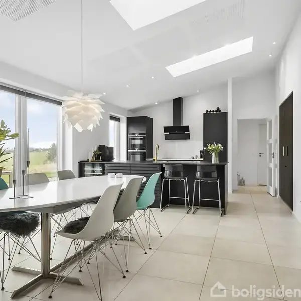 Et moderne køkkenalrum har et langt spisebord omgivet af forskellige stole. Rummet er lyst med store vinduer, et hvidt loft med ovenlys, og sorte skabe og apparater.