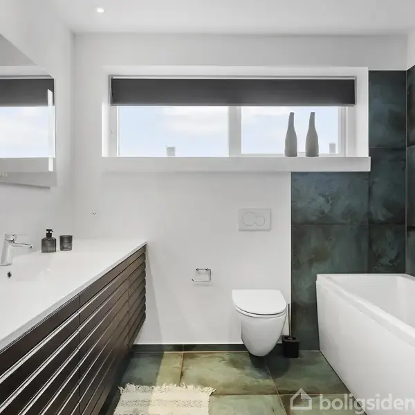 Badeværelse med hvidt badekar og vægmonteret toilet, stående ved siden af en stor kommode med indbygget vask. Store vinduer med persienner giver lys til rummet, der har mørke vægpaneler.