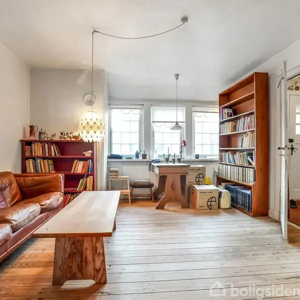 En stue med en brun lædersofa står ved siden af et træbord. Reoler fulde af bøger omkranser rummet. Et spisebord ses nær vinduerne, der tillader naturligt lysindfald.