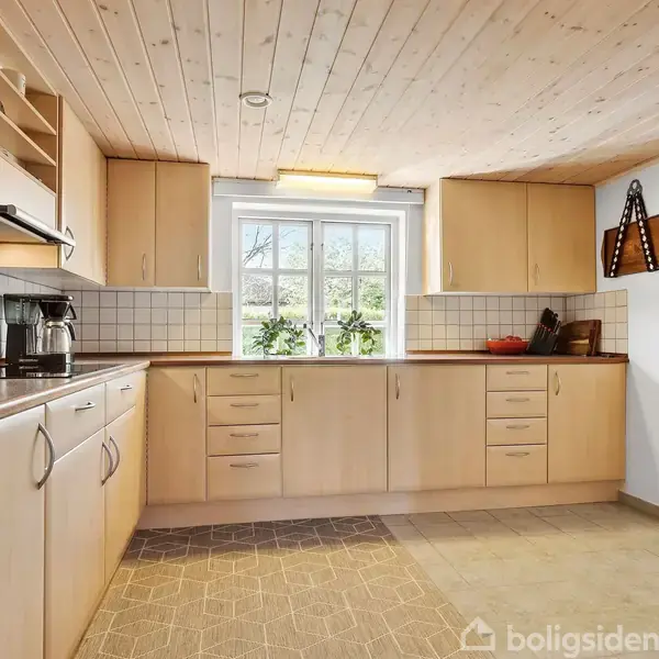 Lyst køkken med lyse træskabe, kaffemaskine og køkkenredskaber på en bordplade, vindue med udsigt til have, flisegulv og træloft. Skærebræt hænger på væggen.