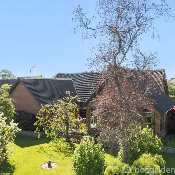 Et rødt murstenshus beliggende i en frodig have, omgivet af grønne buske og træer. Huset har et sort tag, og solen skinner i en klar blå himmel.