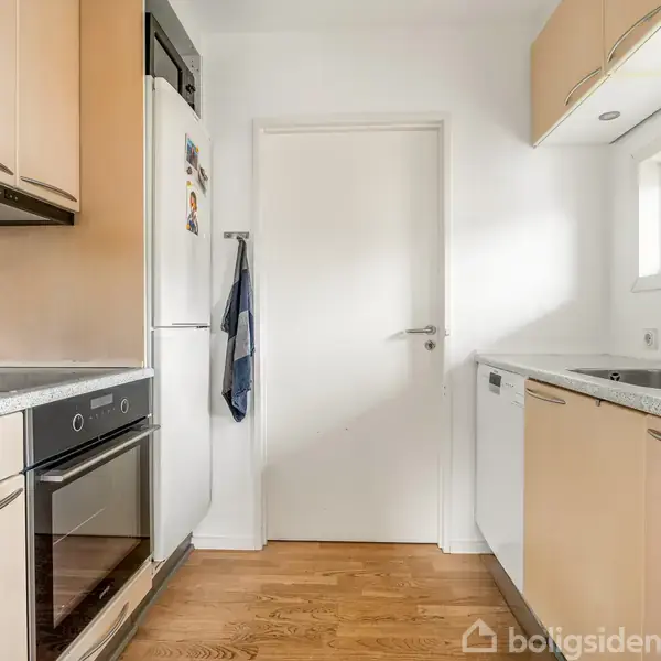Et moderne køkken med lyse trælignende skabe. En ovn og køleskab er integreret, mens en håndvask er placeret ved et lille vindue. Rummet har lyst trægulv og en lukkedør.