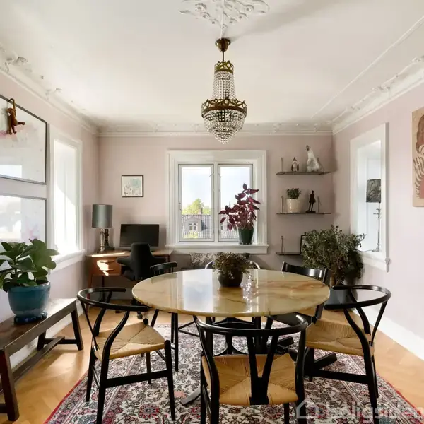 Et rundt bord med stole står i et lysfyldt spiseværelse. En lysekrone hænger over bordet. Væggene har kunst og hylder med pynt. På gulvet ligger et mønstret tæppe.