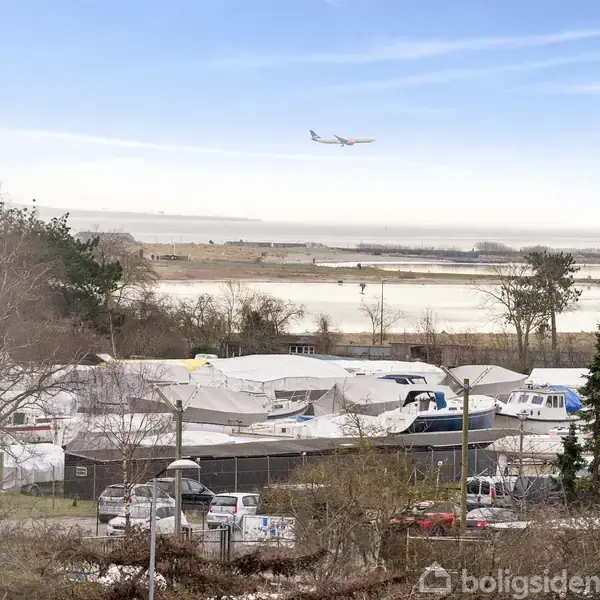 Et fly flyver hen over et kystnært område med træer og parkerede biler samt både dækket af presenninger. Området ligger tæt på en strand og havet kan ses i baggrunden.