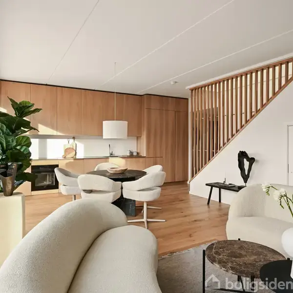 En stue med lys indretning, hvor hvide stole omkranser et rundt bord. I baggrunden ses et køkken med træpaneler og en trappe. En sofa og plante dekorerer rummet.