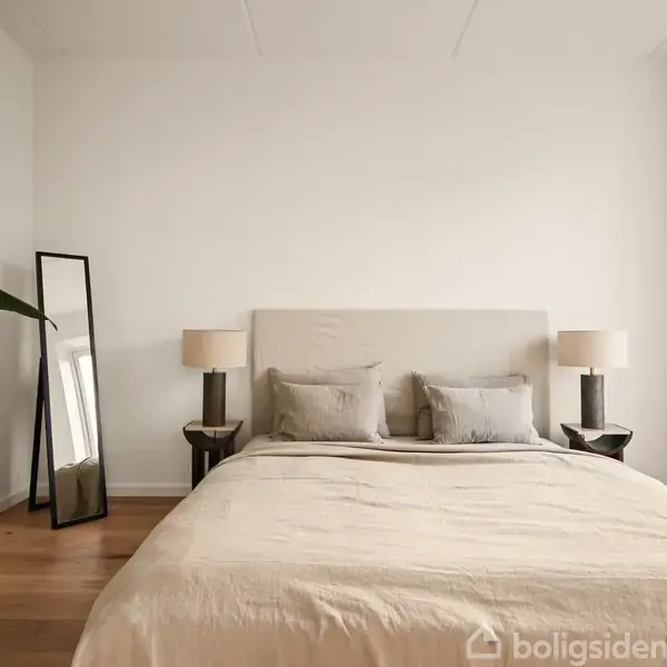 En seng med beige sengetøj er centreret i et soveværelse. To natborde med lamper står på hver side. Et stort spejl og en plante ses til venstre.