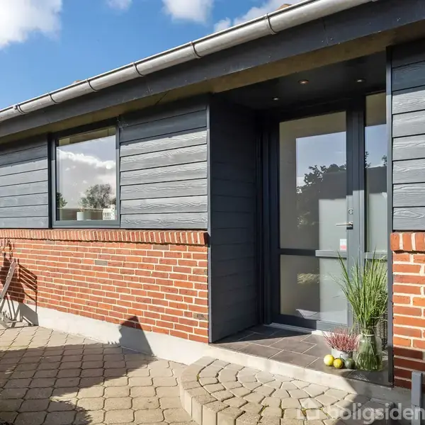 Et hus med murstensfacade og grå beklædning har en moderne hoveddør. Foran døren står to planter i potter. Der er en lille terrasse med fliser, og himlen er blå.