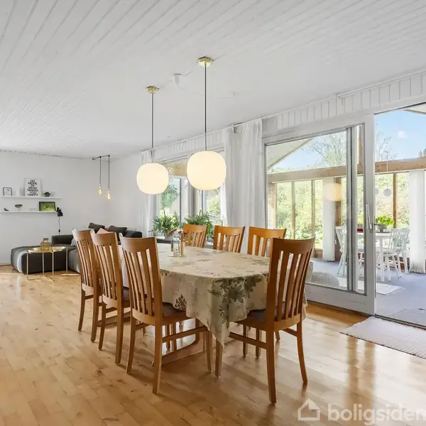 Langbord med træstole står i en rummelig, lys spisestue. To kuglelamper hænger over bordet. Store vinduer viser udsigt til terrasse og grøn have i baggrunden.