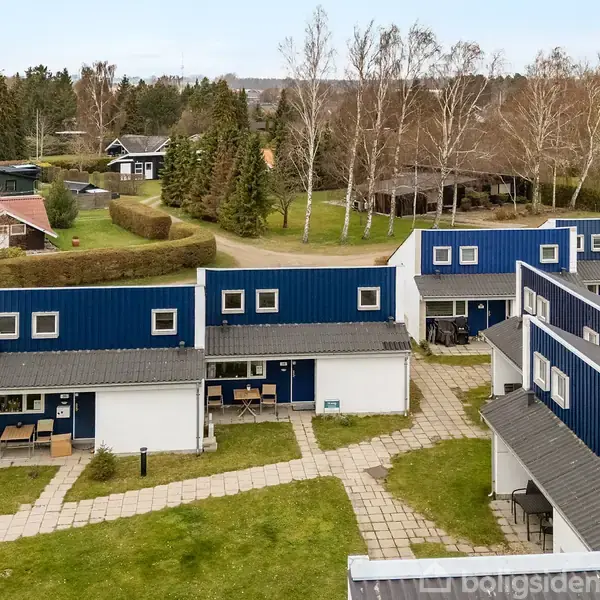 Rækkehuse med blå tage står stillet parallelt. De ligger i et grønt område med stier, omkringliggende træer og flere små huse spredt i baggrunden.