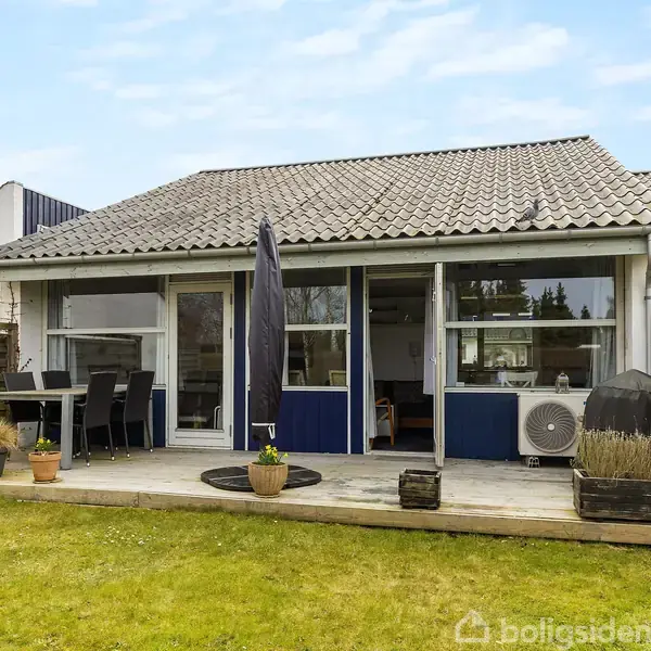 Hus med terrasse, der har havemøbler og en paraply; beliggende i en have med græsplæne og blomsterkrukker. Det omkringliggende miljø er en boligkvarter.