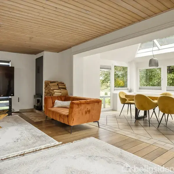 En stue med en orange sofa foran et stort TV, træloft, og trælaminatgulv. Til højre er en spiseplads med stole i gul velour omkring et bord, omgivet af store vinduer.
