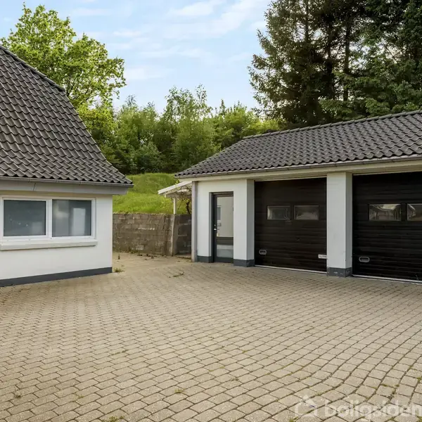 Hus med garage: Huset har hvide vægge og grå flisetag, garage med to sorte porte på en brostensbelagt gårdsplads; omgivet af træer og grønne omgivelser.