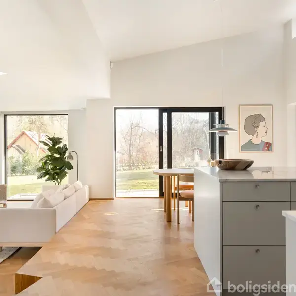 Stue med sofa og plante ved glasvæg, der giver udsigt til have. Et spisebord er placeret ved siden af køkkenøen. Rummets vægge er lyse med et portræt.