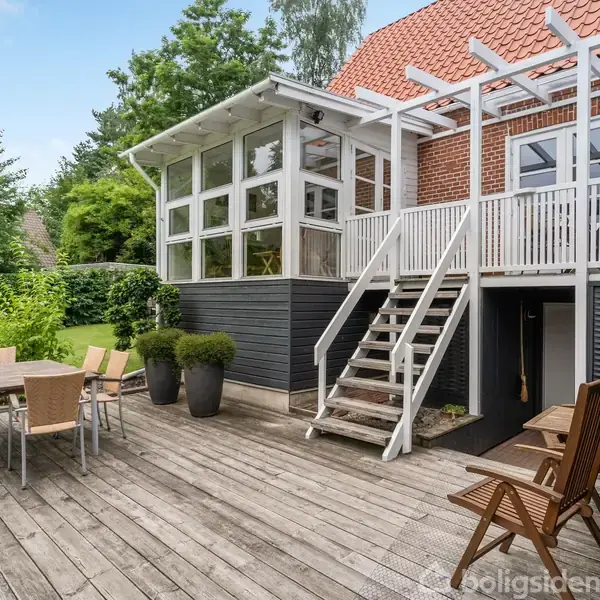 Træterrasse med spisebord og stole foran en rød murstensbygning med hvid trætrappe. Foran er planter og en grøn haveomgivelser. En del af terrassen har tag.