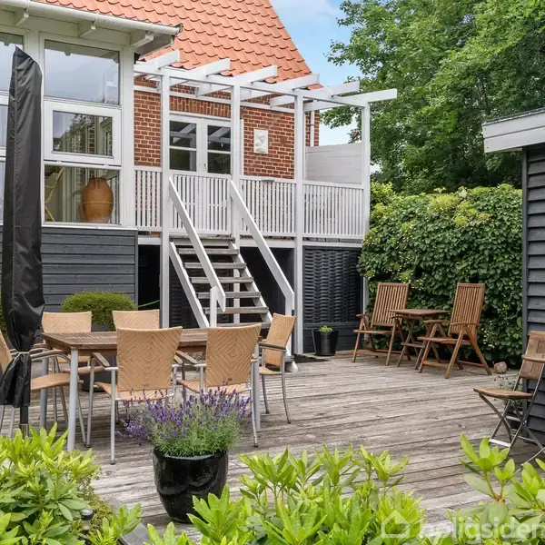 Terrasse med træmøbler og parasol står på et trægulv. En trappe fører op til en bygning med store vinduer. Omgivet af blomster og grønne træer.