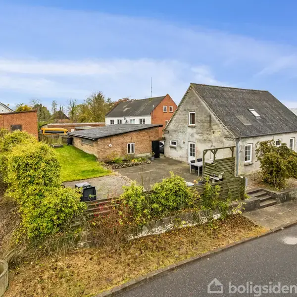 Et ældre hus med en stor tagryg står ved siden af en flisebelagt gårdhave, omkranset af buske og hække. Flere bygninger ses i baggrunden under en blå himmel.