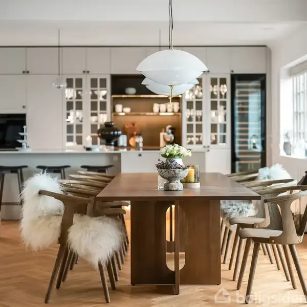 Et træbord omgivet af stole med pels, står i et elegant køkken med lyse skabe og en lampe over. Vindue og dekorationer i baggrunden skaber en lys og indbydende atmosfære.