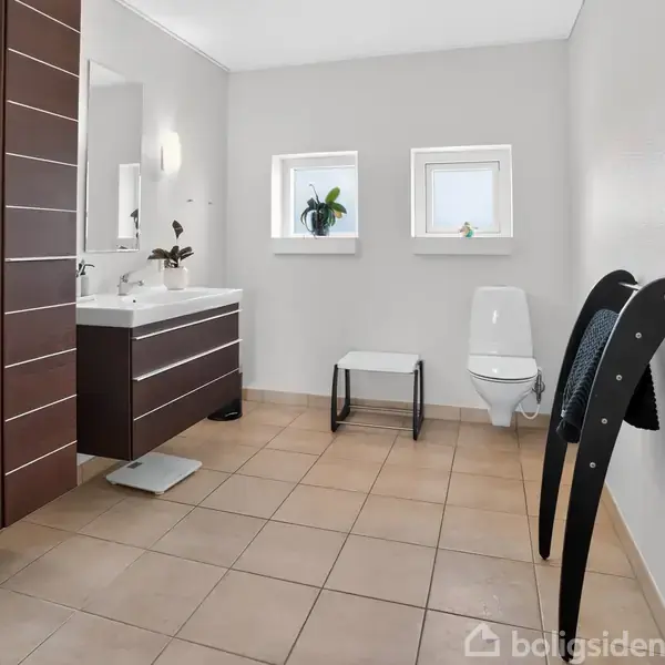 Badeværelse med brun trævask, spejl og håndvask til venstre; hvidt toilet mod væggen; planter og lille bænk under to små vinduer; beige flisegulv.