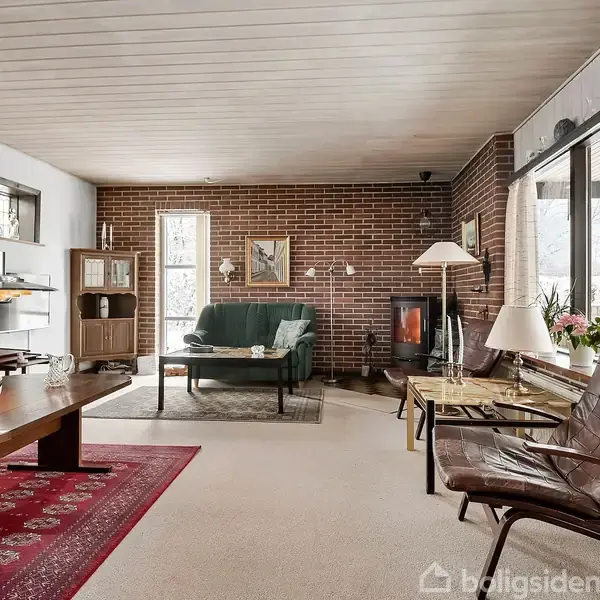 Stue med klassisk indretning, hvor sofa og lænestol står ved et vindue; en brændeovn er på murstensvæggen. Et tæppe og flere lamper skaber en hyggelig atmosfære.